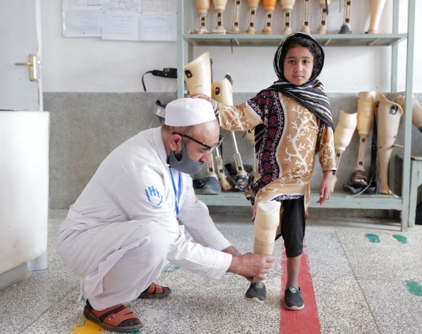 A young Afghan girl balances on one leg while a man in a white lab coat adjusts the artificial limb on her other leg