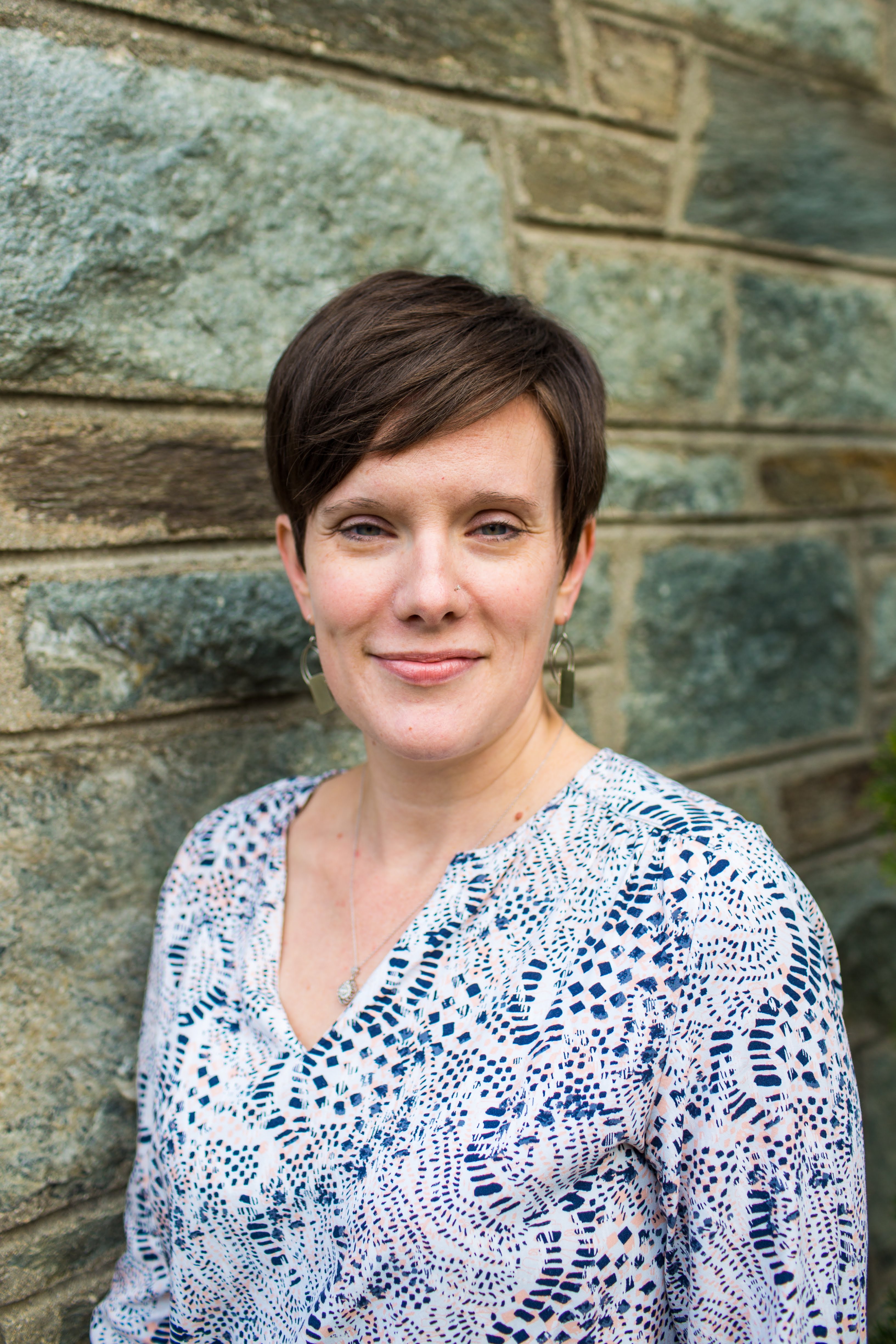 A white woman with short brown hair smiles. She wears a blue and white blouse. She is standing in front of a rock wall.