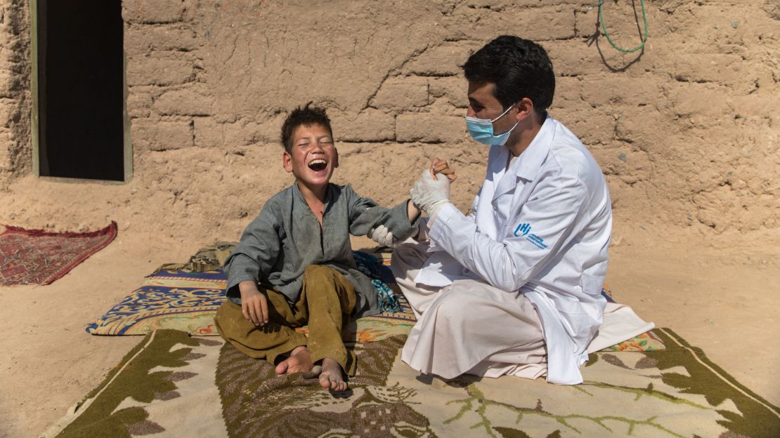 An Afghan boy sits on a blanket smiling. Next to him, a physical therapist in a white coat stretches his arm.