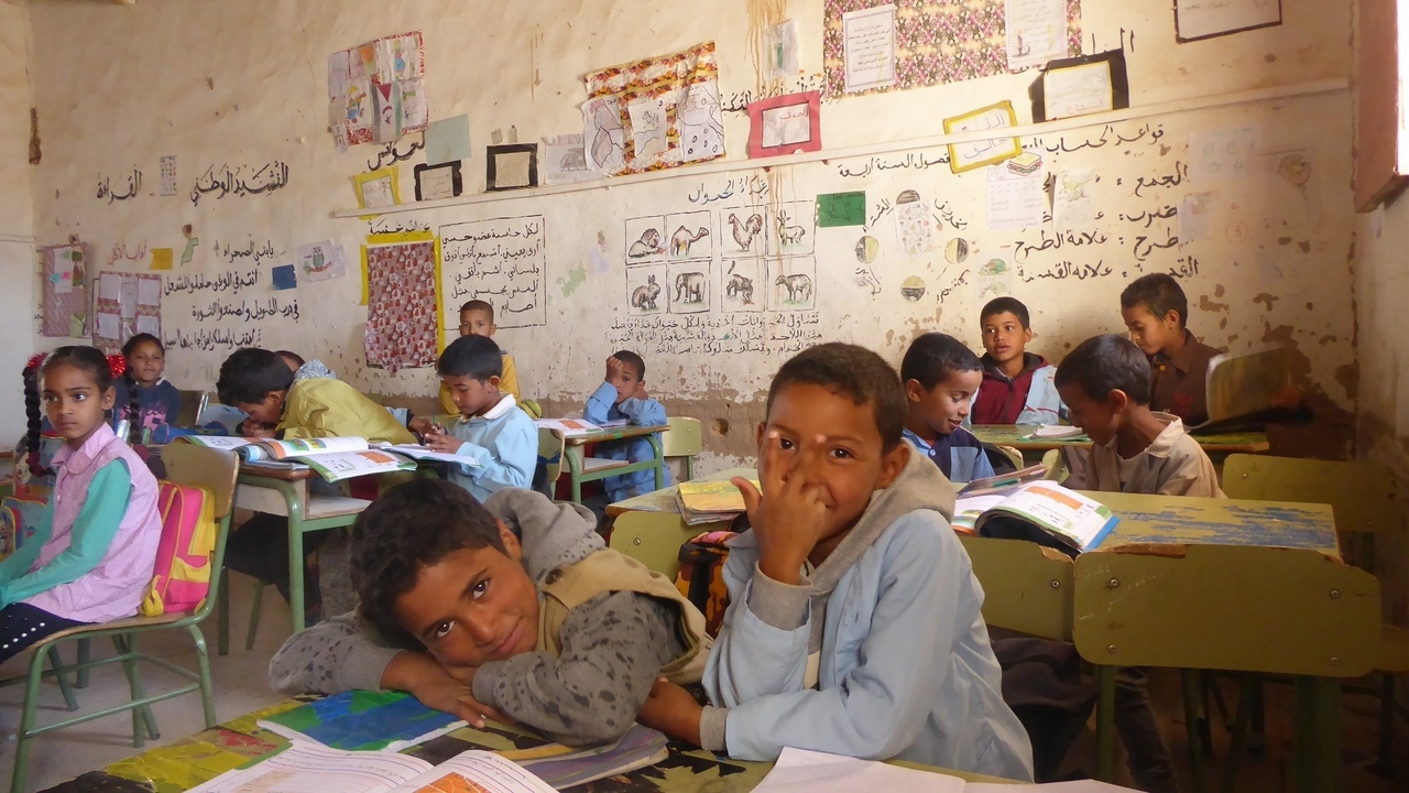 Two young boys smile in a classroom with other children and where the walls are covered in posters.