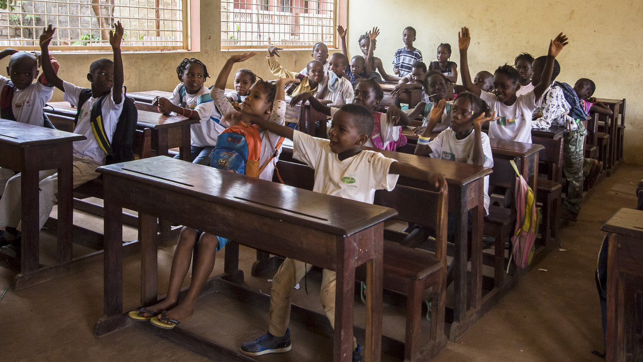 Group of children sitting at desks in a classroom, their hands in the air