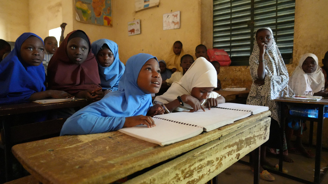 Photo of a classroom in which a girl sitting at a table looks up and smiles.