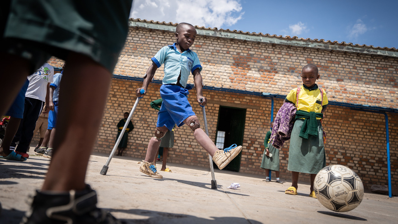 A boy with a prosthetic leg and using crutches is among children playing soccer outside a school.