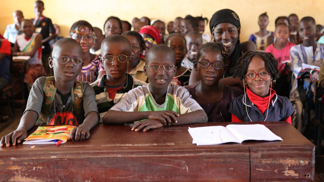 Children, many wearing glasses, smiling as they sit at desks in a classroom.