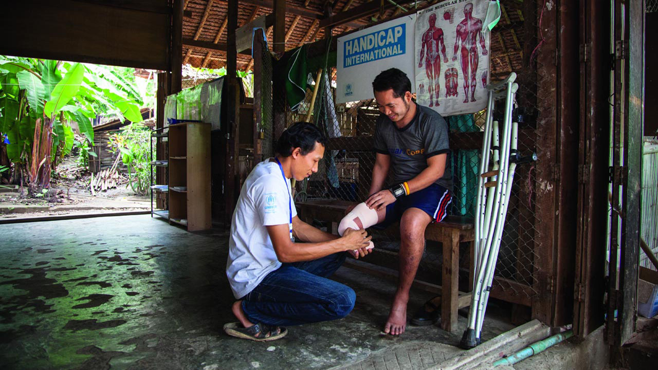A man smiles as his amputated leg is being assessed by an HI worker as he sits on a bench next to crutches. 