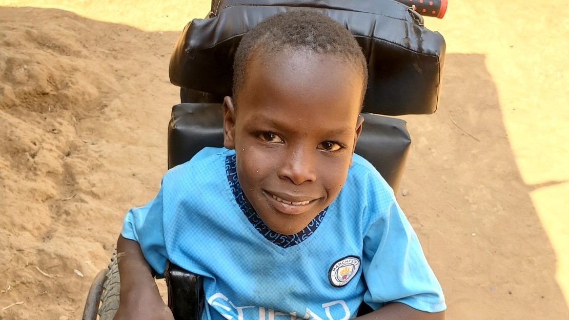 Close-up portrait of a young boy seated in a wheelchair. He looks at the camera and smiles.