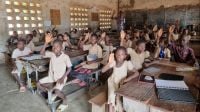Year 2 class taught by Yaovi Ahli at Kombonloaga public primary school in Dapaong, northern Togo. Inside a classroom. Children aged 8–10 in beige uniforms are seated around wooden desks and raising their hands.
