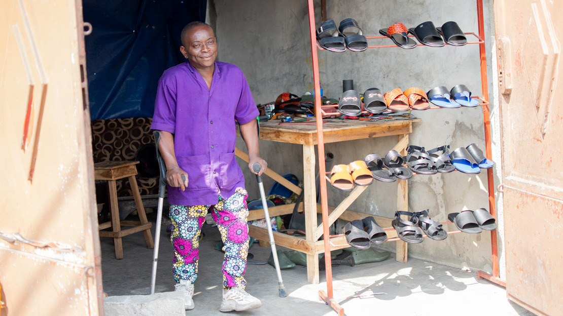 A man stands on crutches between the open doors of a workshop overlooking the street. A display case shows different pairs of sandal-type shoes.