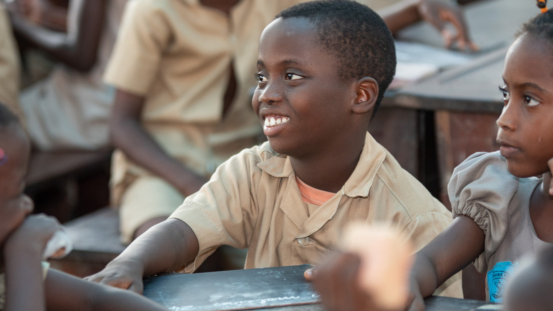 Close-up portrait of a boy sitting in a classroom, holding a slate in his hand. His face is turned three-quarters and he is smiling. Around him, we can make out the faces and arms of other children.