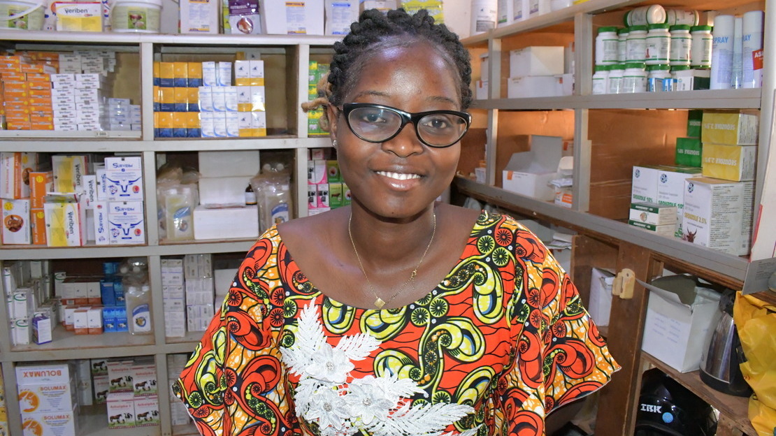 Portrait of a young woman posing in a pharmacy, with shelves full of medicine boxes. The young woman wears glasses and smiles at the camera.