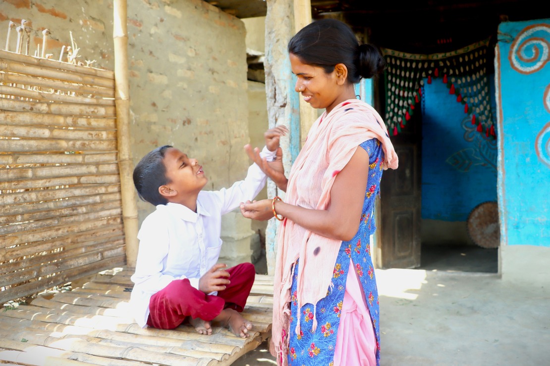 A little boy sits with his arm outstretched towards his mother, who smiles at him in front of their house.