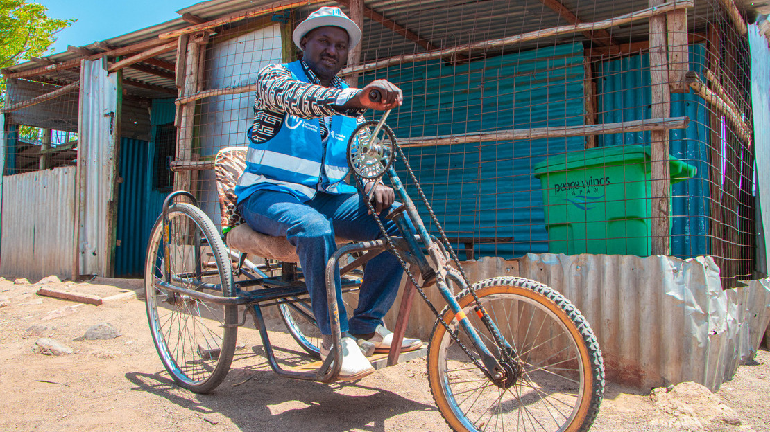 A man sits on a tricycle. He's on a street, and behind him is a tin house. He smiles at the camera.