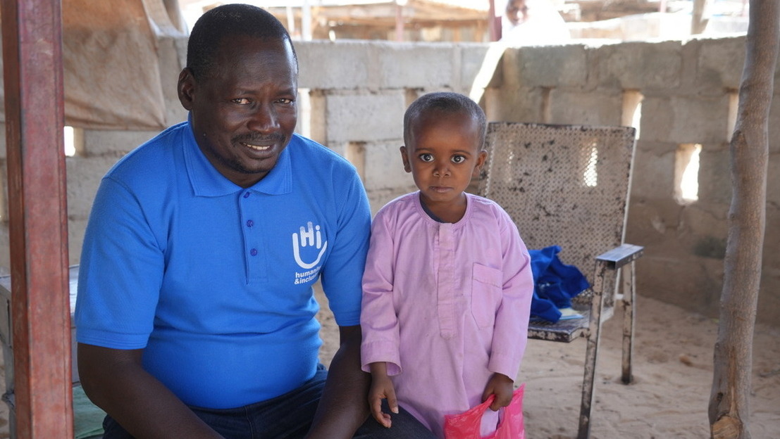 Close-up portrait of a man in a blue HI T-shirt standing next to a boy in a pink jacket. Both smile at the camera.