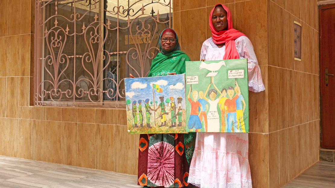 Two women stand in front of a building, holding colorful paintings of fighters shaking hands and children with placards and a white dove.