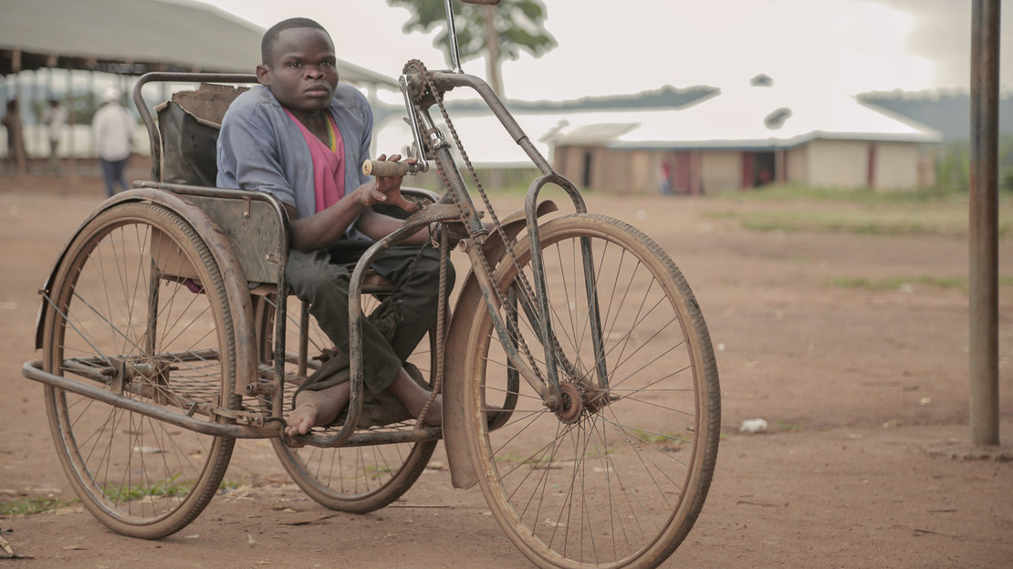 A man sits on a tricycle, looking at the camera. Behind him, we can make out people walking, several buildings and some vegetation.