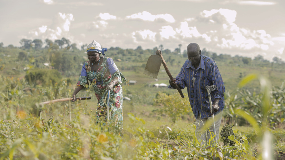 Two people, a woman and a man, are working in a field. Each is holding a spade in their right hand and a crutch in their left.