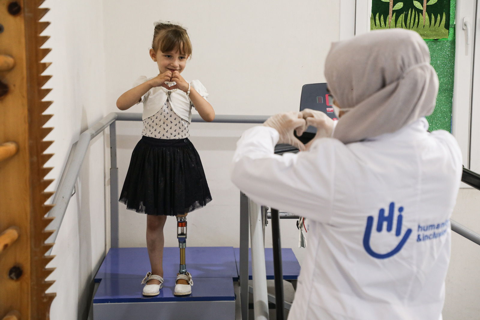 Photo of Enas, a 5-year-old girl wearing a black skirt with a white top and vest. She is standing on a rehabilitation staircase with handrails. You can see her legs, including her prosthetic leg. Enas looks at the HI worker in front of her. Enas and Fatima, the HI worker, make a heart with their hands.