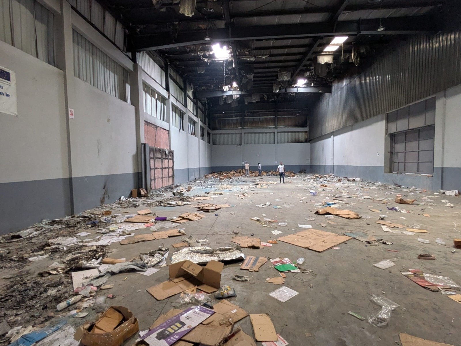 Inside a warehouse: the floor is littered with cardboard boxes, sacks and garbage of all kinds. Part of the roof is black with soot, and sheet metal panels are peeling off.