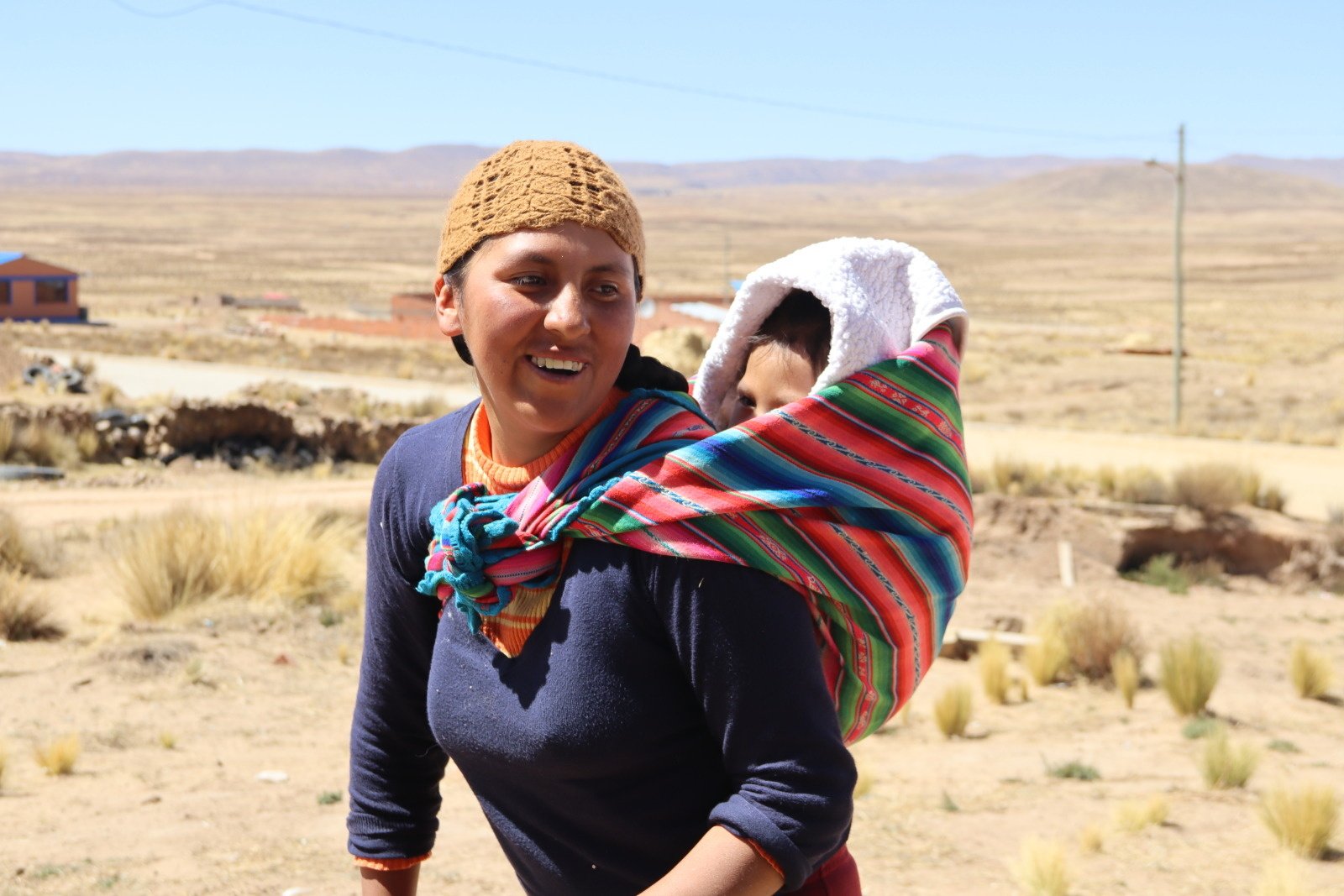 A smiling woman carries her son on her back using traditional aguayo cloth. In the background, the dry, dusty plains of the Altiplano.