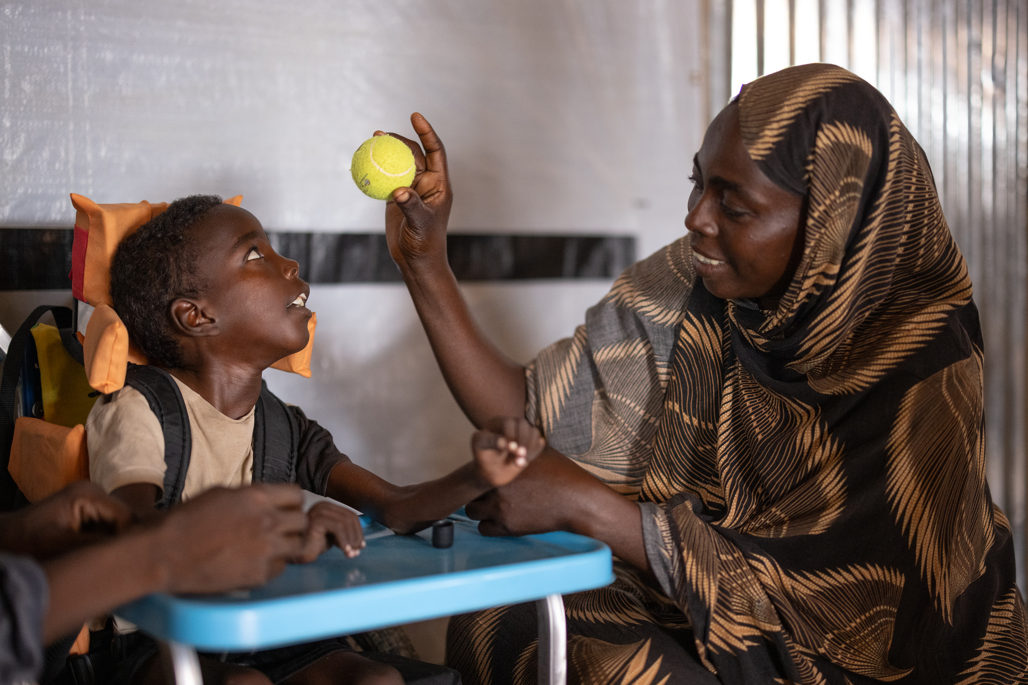 The photo is indoors, in an HI rehabilitation room. Omran, on the left, is in an adapted wheelchair and looks at his mother, who is showing him a tennis ball. Djimilla, his mother, looks at Omran with a smile. 