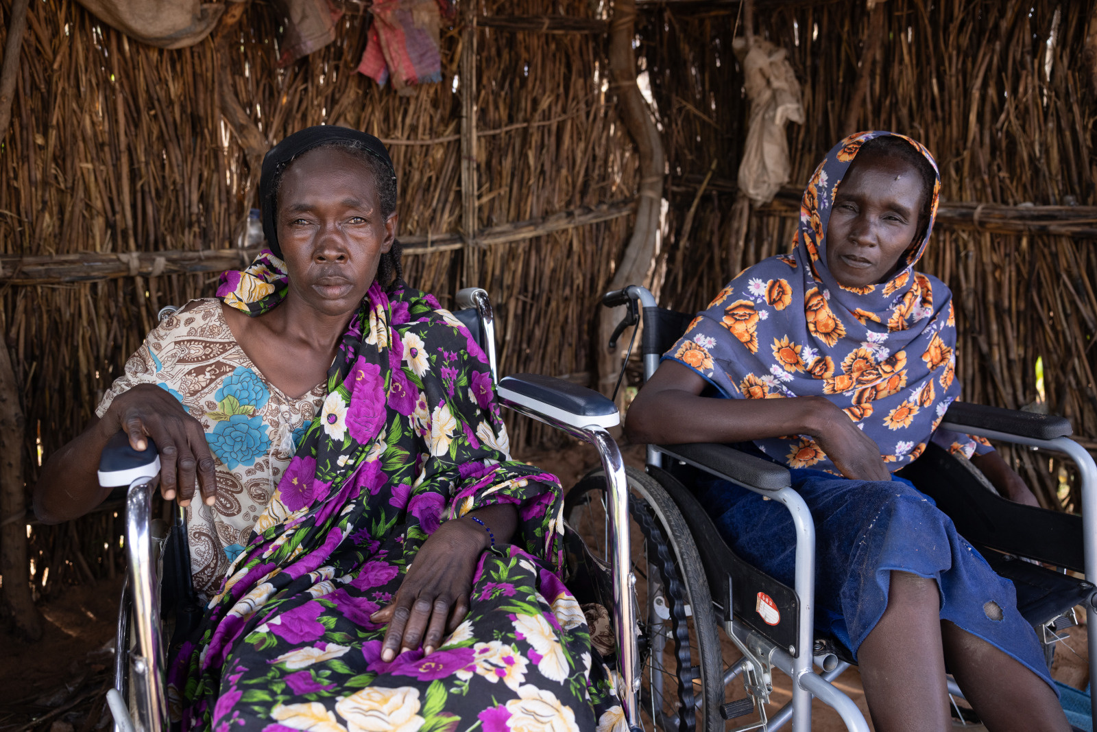 Kaltouma and Hassaneih are side by side, each in a wheelchair. They are sheltered in a wooden house, looking at the lens. Katlouma, on the left, wears a dress with purple and white flowers, while Hassaneih, on the right, wears a blue scarf with orange flowers. 