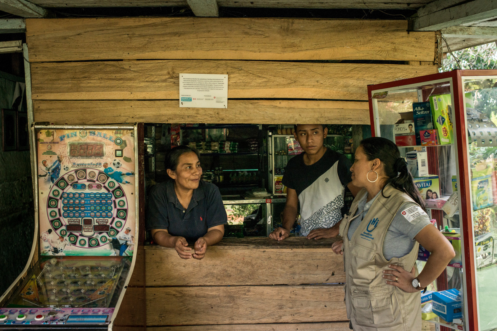 A woman wearing an HI jacket chats with a woman and a man standing behind a wooden counter.