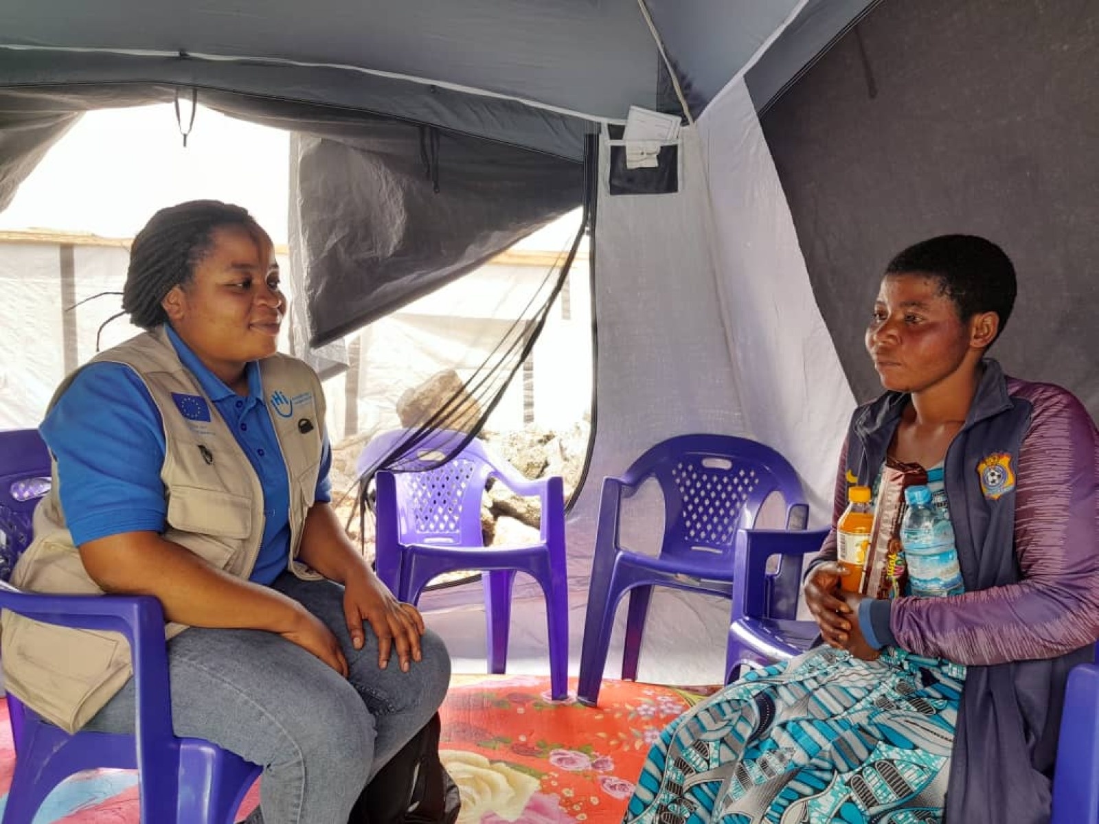 Two women face each other, seated on chairs in a tent. The one on the left is wearing a HI jacket, while the one on the right is holding a bottle of water and some cookies.