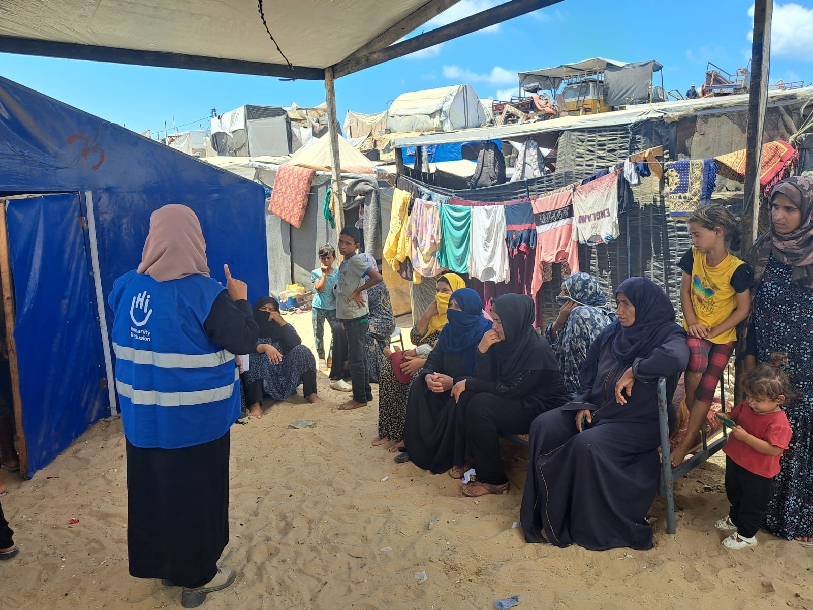 A HI staff woman, wearing a blue vest with the HI logo, and a man, are putting up a sign informing people about explosive remnants. They are in a refugee camp, sticking the sign on a wall. 