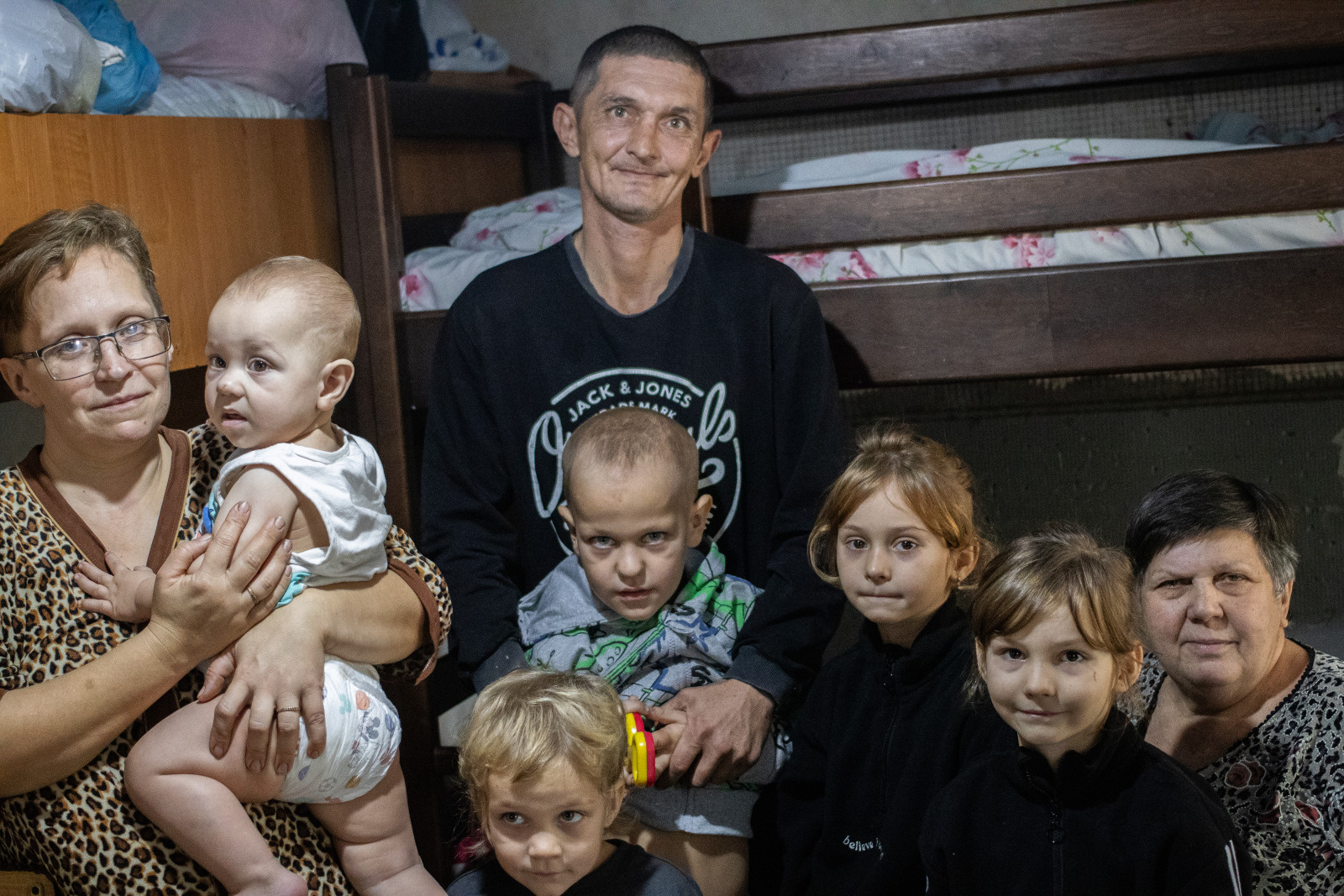 A large family is gathered in a room with bunk beds in the background. The father, in the center, has a gentle smile, while the mother, on the left, holds a baby in her arms. Around them, several children and their grandmother sit or stand, looking at the camera with shy smiles. 