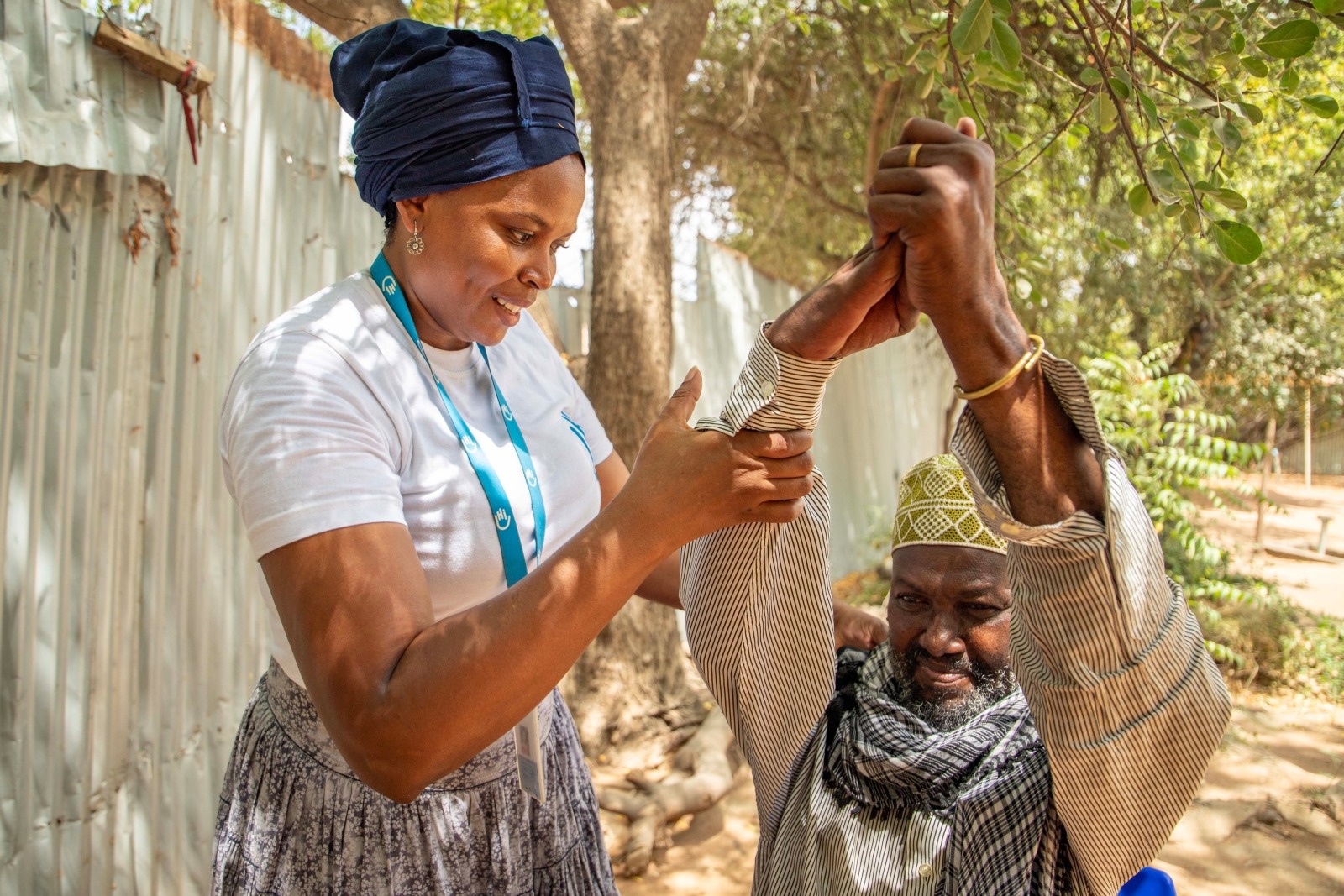 Close-up of a woman holding the raised arms of a seated man. They are in a shady courtyard.