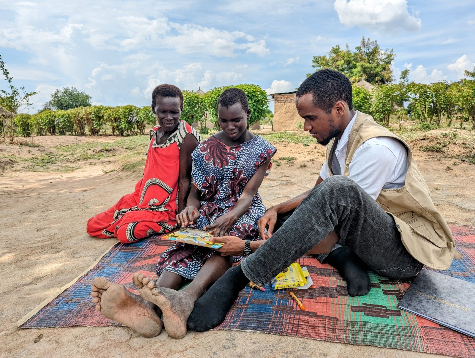 Santina doing a puzzle, accompanied by her mother and an occupational therapist from HI. The three of them are sitting on a carpet outside. Santina is in the center of the picture, and her mother and the occupational therapist, who surround her, watch her doing the puzzle, smiling. 