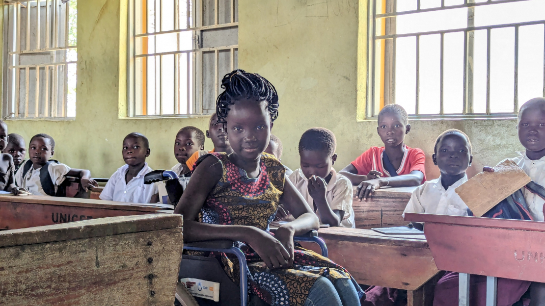 View of a classroom with students seated at desks. In the foreground, a smiling adult woman looks to her left at a young girl in a wheelchair.