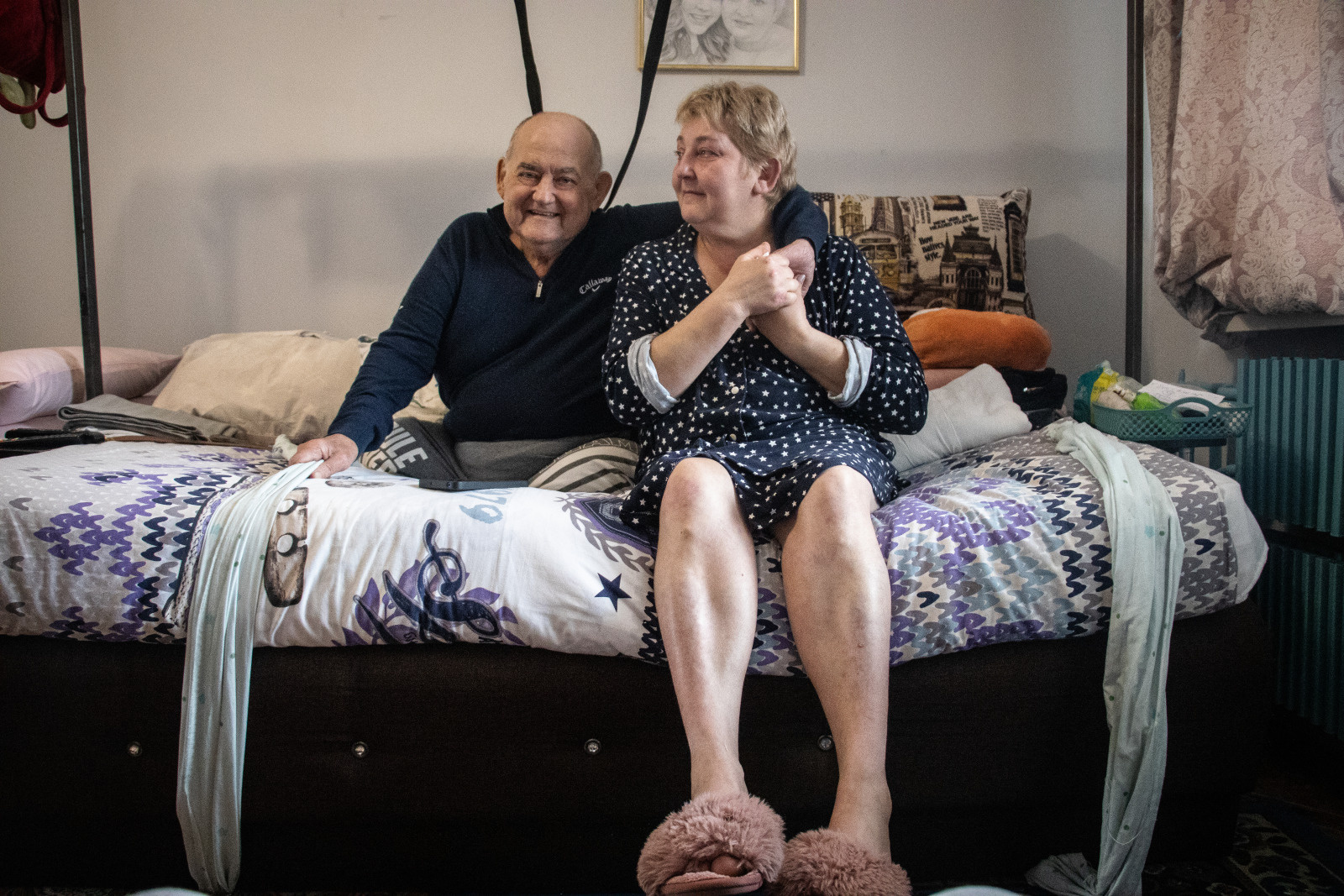 An elderly couple sits on a bed, sharing a moment of closeness. The man, dressed in a dark sweater, smiles warmly, while the woman, wearing a star-patterned robe and pink slippers, gently holds his hand over his shoulder. The intimate atmosphere of the room, with its colorful bedding and everyday objects, creates a feeling of warmth and connection.