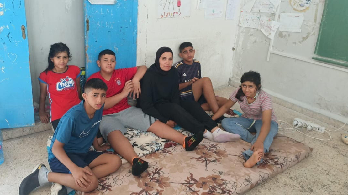 A woman and her five children sit on mattress pads in a classroom.